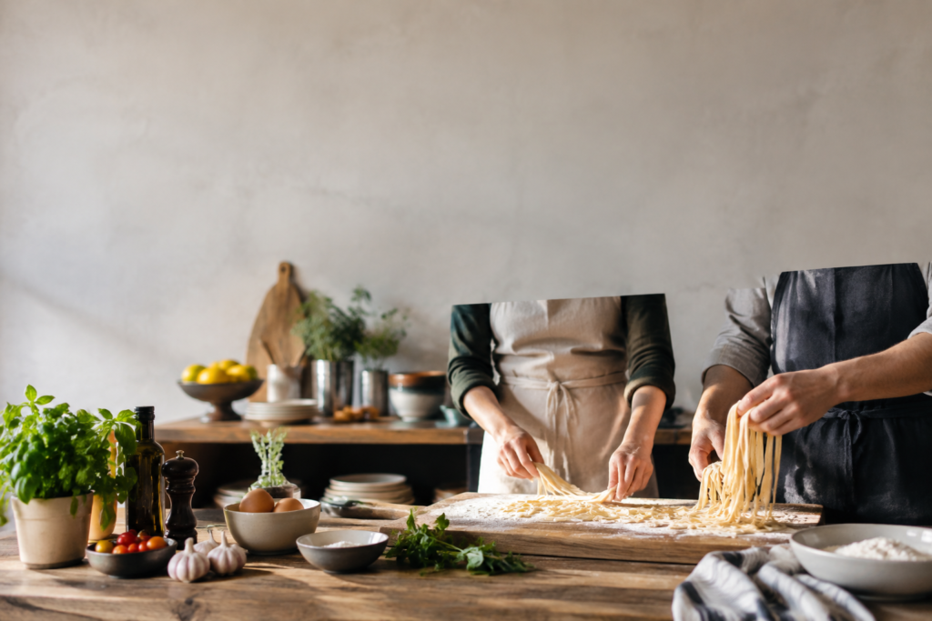 Deux personnes en tablier préparant des pâtes fraîches artisanales sur une planche en bois dans une cuisine lumineuse.