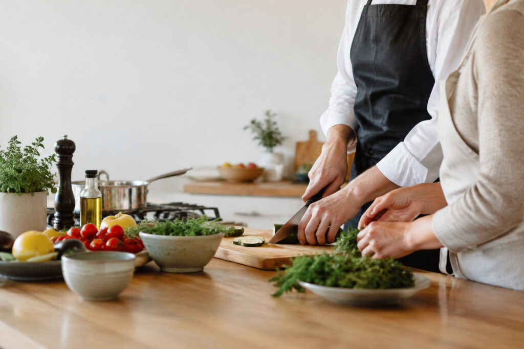 Un chef et son élève coupent des légumes frais sur une planche en bois dans une cuisine moderne et lumineuse.