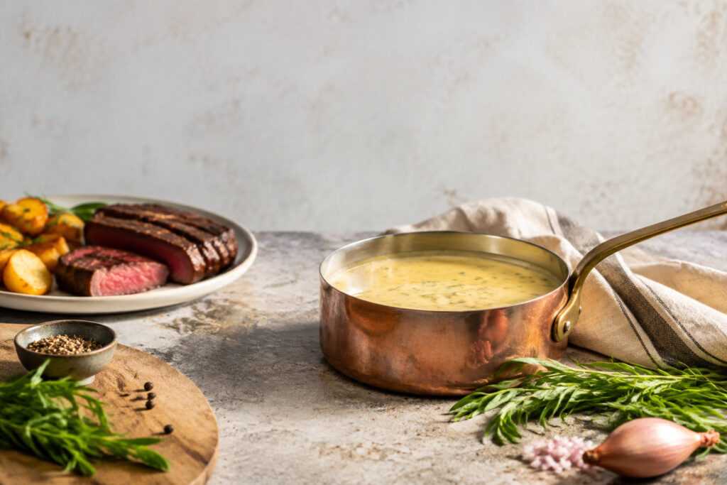 Casserole en cuivre avec une sauce béarnaise, accompagnée de bœuf grillé et d'herbes fraîches sur une table en pierre.